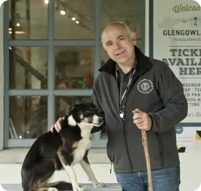 A man in a grey jacket stands beside a black and white dog, gently resting his hand on the dog's back. They are in front of a building with a sign that reads "Tickets Available Here.