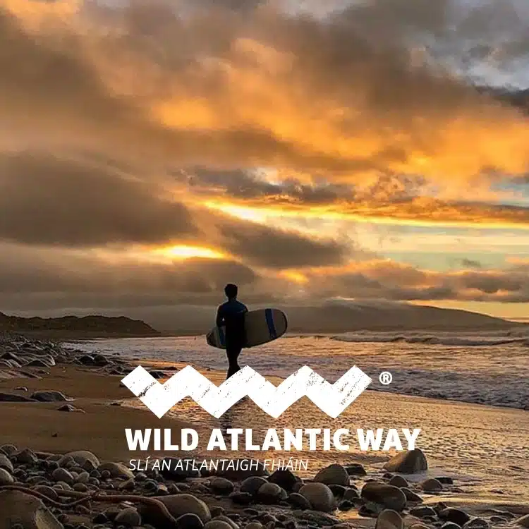 A person with a surfboard stands on a rocky beach at sunset, with dramatic orange and yellow clouds over the sea. Text reads: "Wild Atlantic Way Sli An Atlantaigh Fhiain.