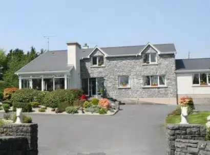 A two-storey house with a stone facade, white trim, and a well-maintained garden in front. There are two white statues at the entrance next to a paved driveway. The sky is clear and sunny.