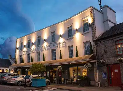 The Connemara Lake Hotel is lit up at dusk, with several cars parked in front and hanging plants decorating the entrance. The white facade and large windows stand out against a partly cloudy evening sky.