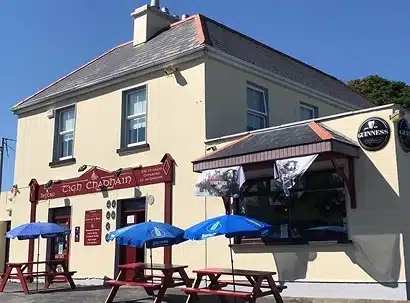 A cream-coloured two-storey pub with red trim and signs reading "Tigh Chadhain." Blue picnic tables with umbrellas are outside, and Guinness signs hang above the entrance. The sky is clear and sunny.