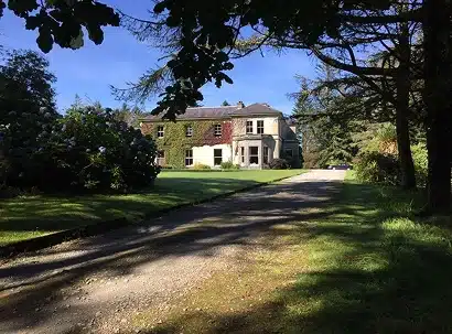 A large country house with ivy-covered walls sits at the end of a gravel drive, surrounded by green lawns, trees, and shrubs on a sunny day.