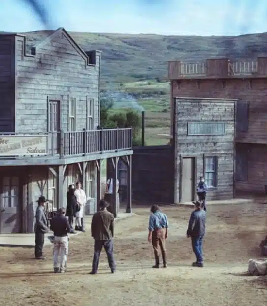 A group of men in cowboy attire face each other on a dirt street in a Western-style town with wooden buildings and a hilly landscape in the background.