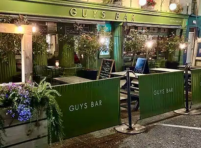 Outdoor seating area of Guys Bar, with green barriers, wooden tables, potted plants, fairy lights, and a chalkboard menu in front of a green-painted building at night.