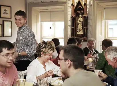 A group of people are dining together in a cosy restaurant. A smiling waiter stands near the table, while guests eat, talk, and enjoy their meals. The atmosphere appears friendly and lively.