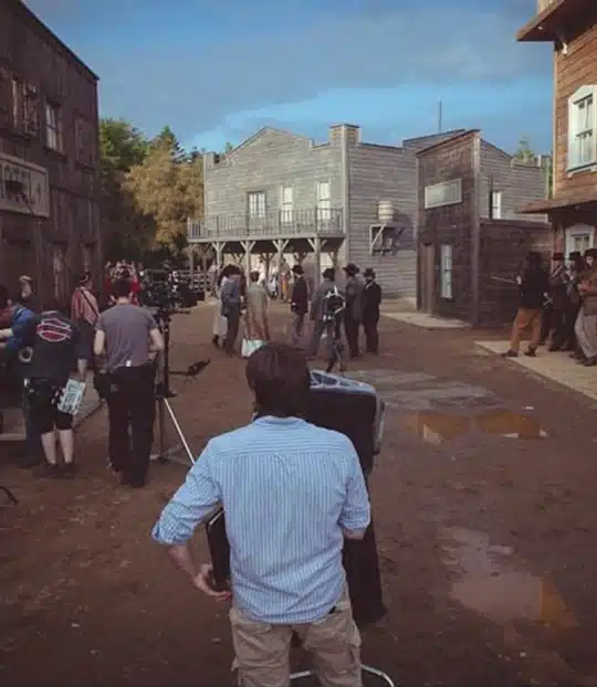 A film crew sets up equipment on a muddy street of a Western-style film set, with actors in period costumes and wooden buildings in the background under a partly cloudy sky.