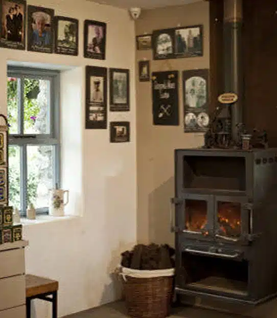 A cosy corner with a wood-burning stove, a basket of peat, framed photos on cream-coloured walls, a window with light coming through, and shelves with tins and a mug on the windowsill.