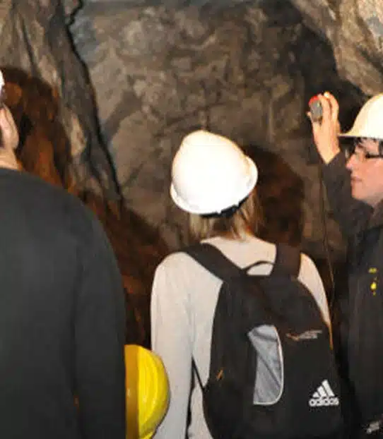 Several people wearing hard hats explore a rocky cave or mine; one person holds up a torch or device, and another carries a black rucksack with an Adidas logo.