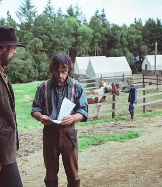 A man in braces reads a paper while standing next to another man in a hat on a dirt path. Behind them are wooden fences, horses, a person in the distance, and tents with trees in the background.