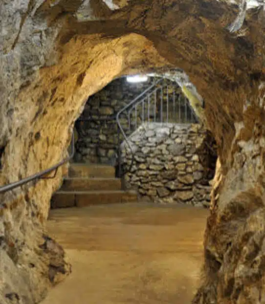 A stone tunnel with rough, uneven walls and ceiling leads to a staircase of stone steps with a metal handrail, ascending towards a lit area with a stone wall in the background.