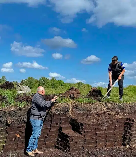 Two people work outdoors on a peat bog under a blue sky with scattered clouds; one stands on a lower level while the other uses a long tool on the upper layer of cut peat blocks.