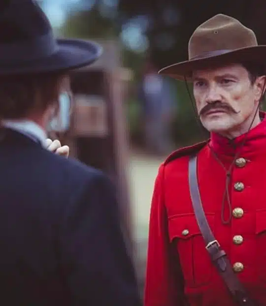 A man in a red uniform and brown hat, resembling an historical Mountie, stands outdoors facing another person in dark clothing and a wide-brimmed hat. The background is blurred with greenery.