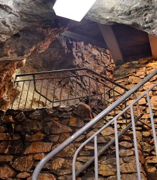A stone staircase with a metal bannister leads upward through rough, uneven rock walls inside a dimly lit stone structure, with a ceiling partially lined with wooden beams.
