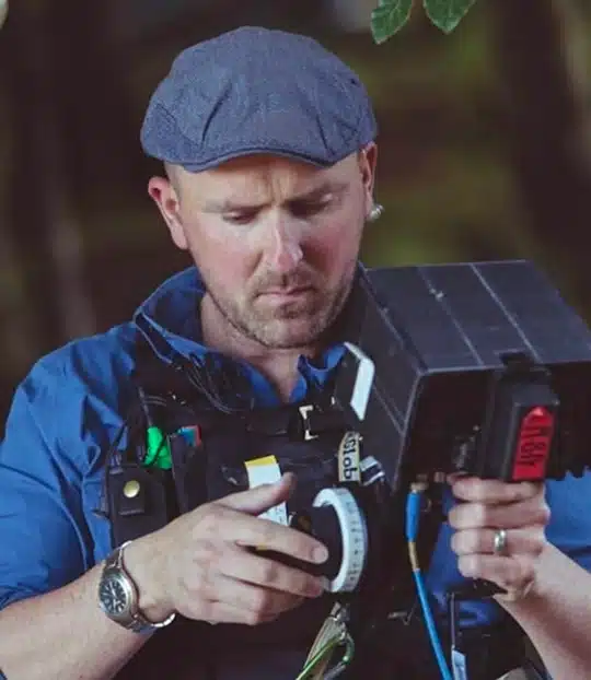 A man wearing a flat cap and blue shirt operates a professional video camera outdoors, adjusting settings whilst focusing on his work.