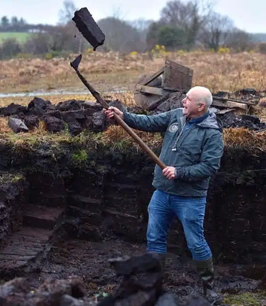 A man wearing a raincoat and wellies stands in a muddy field, tossing a lump of peat into the air with a long-handled spade. The background shows grassy, rural countryside with trees and shrubs.