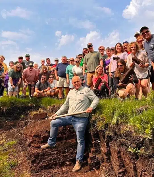 A group of people pose outdoors on a grassy hill under a blue sky. One person sits in front on a ledge holding a long tool, whilst the rest stand and smile behind him. It appears to be a group event or tour.