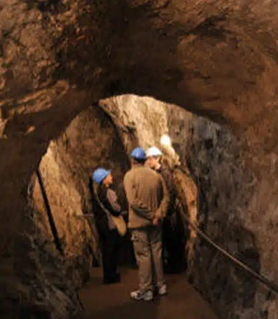 Three people wearing blue hard hats stand and talk inside a dimly lit, narrow, rocky tunnel. The rough walls and ceiling suggest it is an underground cave or mine. A handrail runs along one side.